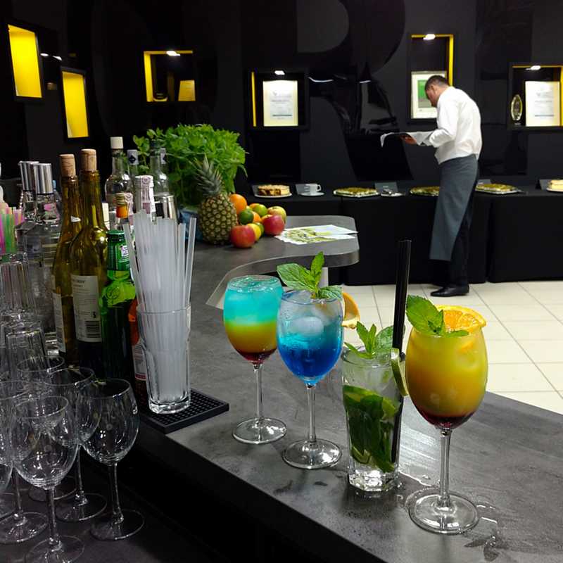 Koktajle w kieliszkach stoją na mobilnym barze. Barman, butelki, owoce cytrusowe i zioła, czarne ściany.